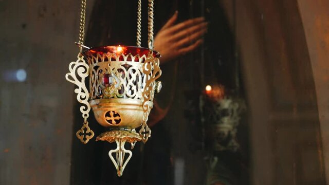 Hanging icon lamp of transparent red glass in golden holder in Orthodox church. Gilded church altar utensil. Closeup. burning light in an old copper church lamp. In the background is an image of a
