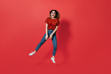 Fototapeta na wymiar Full-length portrait of perky woman in jeans and glasses jumping on red background