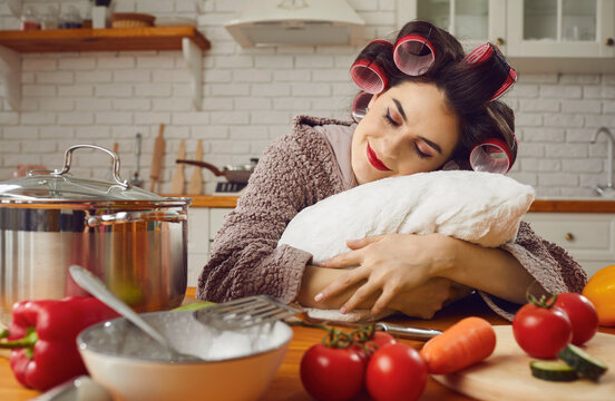Happy funny lazy housewife taking a nap amid cooking a meal. Tired pretty young woman in curlers and bathrobe sitting at a kitchen table, hugging a pillow, sleeping and dreaming sweet peaceful dreams