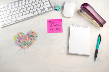 Words Happy Valentine's Day in a note on the desktop with colored paper clips in the shape of a heart and keyboard, pen, notepad and stapler at workplace. Valentines day in office.