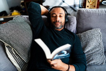 Handsome man relaxing on the sofa at home with a book and headphones