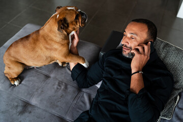 Happy mixed race man playing with his bulldog on the couch at home and talking on the phone