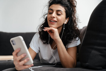 Pleased woman in earphones using mobile phone while lying on sofa