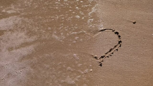 Woman draws heart on a wet sand. Female's hands writing the word sea on sand. Sea water washes away the drawing on a sandy beach.