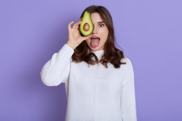 Young adorable european female in white casual clothing covering her eye with half of avocado, keeps healthy eating, looks at camera and showing tongue, poses isolated over lilac background.