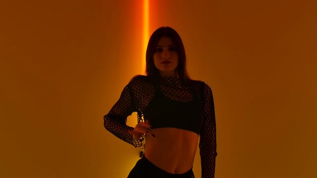 Sexy stripper erotically dances strip with elements of aerobics in dark studio against background of yellow red neon lights. Hot brunette plastic moves demonstrating her body. Close up. Slow motion.