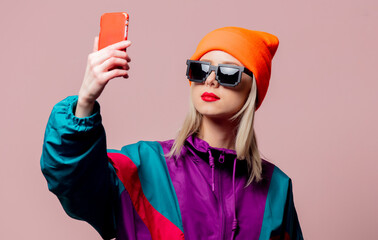 Style girl in 80s sportsuit and sunglasses hold mobile phone on pink background