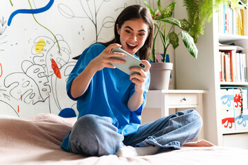 Excited girl playing online game on mobile phone while sitting on bed © Drobot Dean