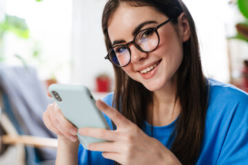 Happy brunette girl in eyeglasses using smartphone and smiling at home
