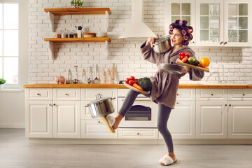 Tired housewife cooking food and carrying lots of stuff. Frail slim young woman making meal at home, holding multiple heavy cooking pots and kitchen saucepans, balancing cutting boards with vegetables
