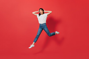 Emotional Asian woman in jeans jumping on red background