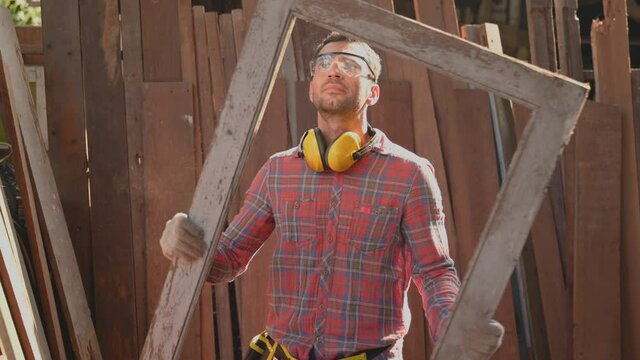 Carpenter young man wearing safety goggles standing with checking perfection of old window jamb for repair in workplace at furniture factory. Concept of small business owner and startup about woodwork