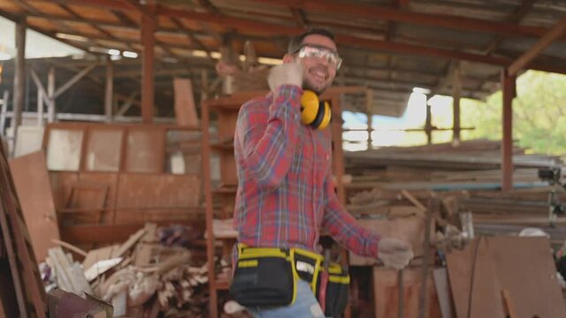 Portrait of happy carpenter young man wearing plaid shirt standing smile and dance after working completed in workplace at furniture factory. Concept of small business owner and startup about woodwork
