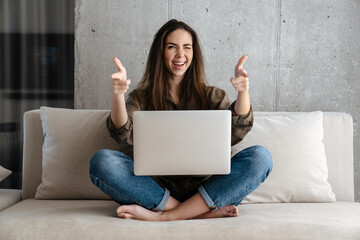 Joyful girl winking and pointing fingers at camera while using laptop