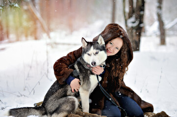 Portrait of a beautiful girl on a walk on a Sunny winter day with a Husky dog in the Park. The girl gently hugs the dog and looks at the camera. A man on white snow with a dog. People and dogs.