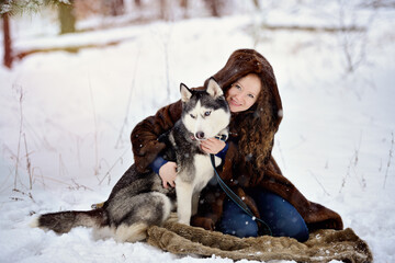 Portrait of a beautiful girl in a fur hood on a winter day with a Husky dog in the Park. The girl gently hugs the dog and looks at the camera. On white snow with a dog. People and dogs.