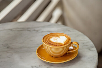 Trendy yellow cup of hot cappuccino on marble table background. Heart shape latte art for symbol of love