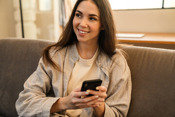 Happy young woman smiling and using smartphone while sitting on sofa