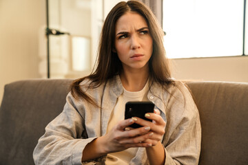 Displeased young woman using smartphone while sitting on sofa indoors