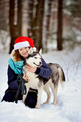 Fototapeta na wymiar Southern smiling girl in a Christmas hat with a Husky dog in the winter forest. She sits and hugs her dog. She looks into the camera lens. Vertical orientation.