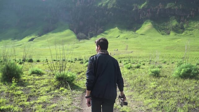 Man walking on beautiful meadow landscape at foot of Bromo Mountain National Park at Teletubbies hill (Bukit Teletubbies Bromo) and holding a camera, at East Java, Indonesia. Wide shot.