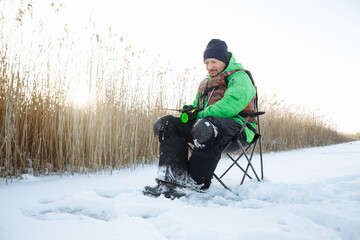 Fototapeta na wymiar a young man on a winter fisherman sits on a chair and fishes and a fishing rod