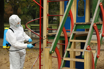 Fototapeta na wymiar Woman wearing chemical protective suit with disinfectant sprayer on playground. Preventive measure during coronavirus pandemic