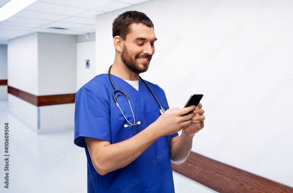 Wall mural medicine, healthcare and technology concept - happy smiling doctor or male nurse in blue uniform with stethoscope using smartphone over hospital corridor on background