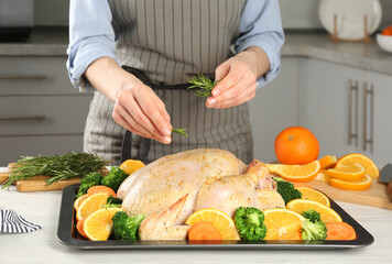 Woman adding rosemary to chicken with oranges and vegetables at table in kitchen, closeup