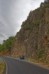 A Black Fiat Car parked in the layby of a Small Spanish Mountain Road, with two cyclists riding up the gentle hill between the Cliff walls.
