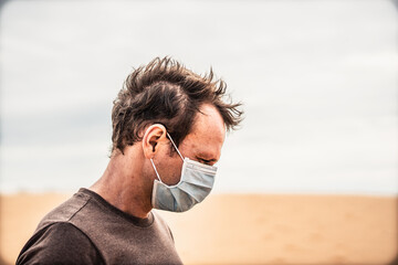 Side portrait young man wear protective mask, face expression emotion look down dune. Concept COVID stress syndrome, mental health, fear of danger contamination, adverse socio economic consequence