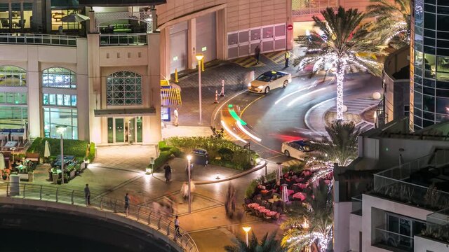 Aerial top view at night timelapse of Dubai Marina promenade with restaurants and canal with floating yachts and boats in Dubai, UAE. Illuminated modern towers with blinking lights and traffic on
