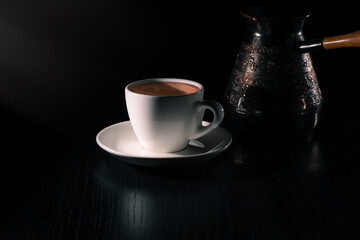 White cup of fresh hot coffee and copper cezve on black wooden table in the rays of morning light. Low key. Copy space.