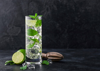 Highball glass of Mojito cocktail with ice cubes,mint and lime on black board with wooden squeezer and fresh limes.