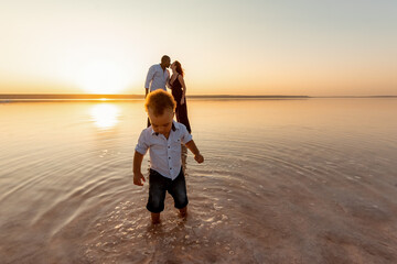 Portrait of little mixed race boy. Kissing parents on background. Happy multi ethnic family at the beach. Beautiful sunset