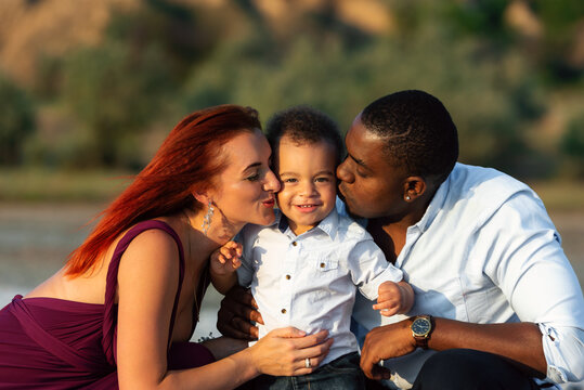 Family closeness. Happy days filled with fun. Parents with son on the beach. Mom and dad kissing their biracial son. Mixed race family