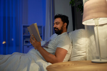 people, bedtime and rest concept - indian man reading book in bed at home at night