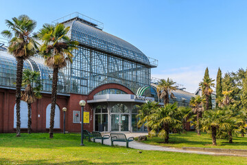 Buildings of palm garden in Frankfurt. Palm trees with meadow in front of the entrance. Glass roof with windows and bricks from the building. Path with benches and lamps - obrazy, fototapety, plakaty