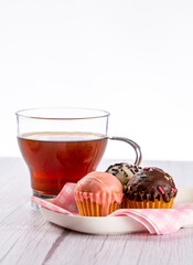 Cup of tea on a wooden table in light tone and white background, accompanied by a plate with mini cup cakes covered in strawberry, dark chocolate and white chocolate.