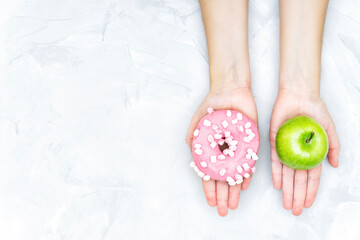 Obraz na płótnie Canvas Crop view of female hands holding a fresh apple and a pink donut offering to make a choice. Healthy eating balance concept.