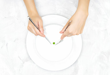 Female hands trying to catch a small green pea on a large white plate with knife and fork. Eating disorder concept.