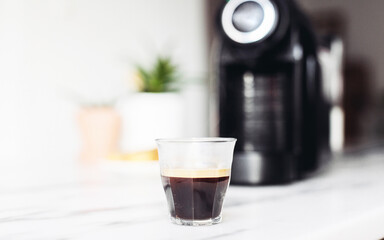 A glass of espresso, black coffee machine on a marble table, white kitchen 