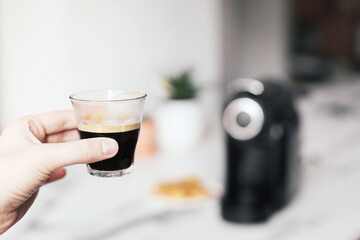 Holding a glass of espresso. Black coffee machine, bread, marble table on the background 