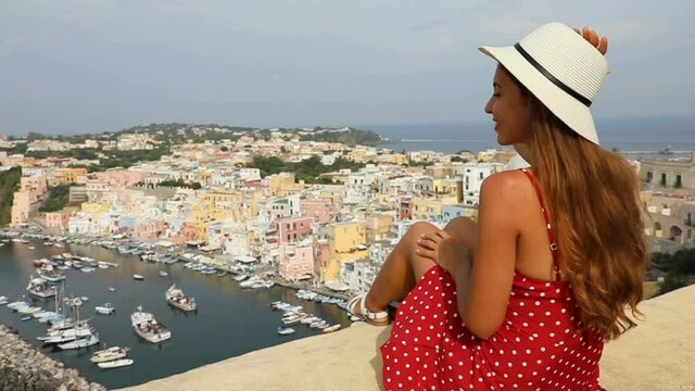 Beautiful young woman with hat sitting on wall looking at stunning panoramic of Procida Island on Naples Gulf, Italy. Slow motion of beautiful girl in Procida, Italian Capital of Culture 2022.