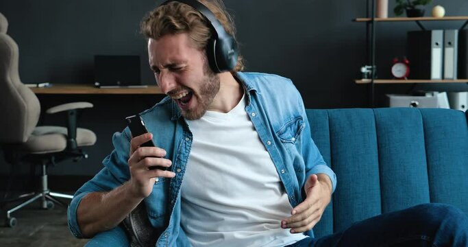 Caucasian Bearded Hipster on the bed and listening to Music in Headphones. Young Man enjoying cool Music on Couch in Loft Living Room. Playfully Mood at Home. FunnyMan. Hobby. Apps.