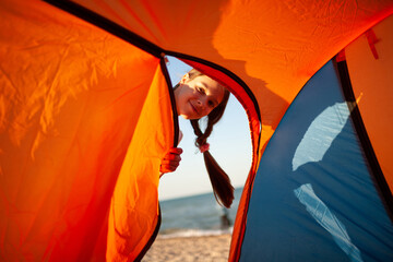 Happy beautiful cheerful young girl stands near a bright tent on the sandy shore of the blue sea and smiles looking at the camera