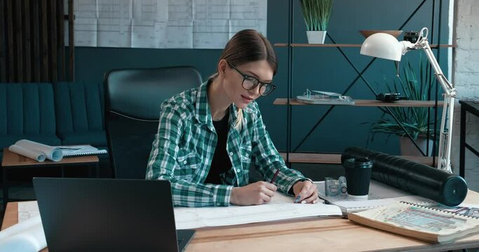 Attractive Woman Architect Making drawings with pencil in Modern Office. Looking at the camera with a Charming Smile in trendy eyeglasses and dressed in Shirt. Portrait.