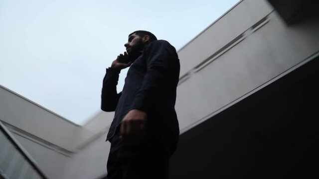 Oriental Man with Beard in a Black Shirt Speaks on Mobile Phone near the Business Center. Serious Guy Talking on Smartphone in the Downtown. Bottom view. The camera rotates quickly 360 degrees.