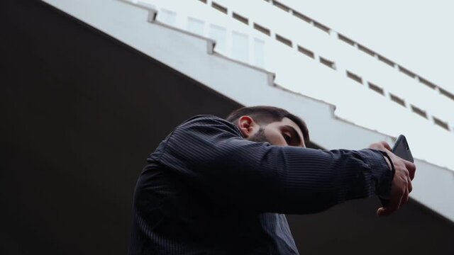 Oriental Man with Beard in a Black Shirt Speaks on Mobile Phone near Business Center. Serious Guy Talking on Smartphone in Downtown. Bottom view. The camera rotates quickly 360 degrees. Slow motion.