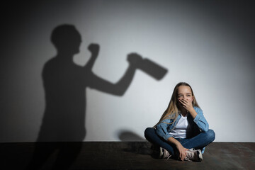 Domestic physical violence, abusing. Scared little caucasian girl, victim sitting close to white wall with shadow of angry threatening father with alcohol addiction. Awareness of social problem.